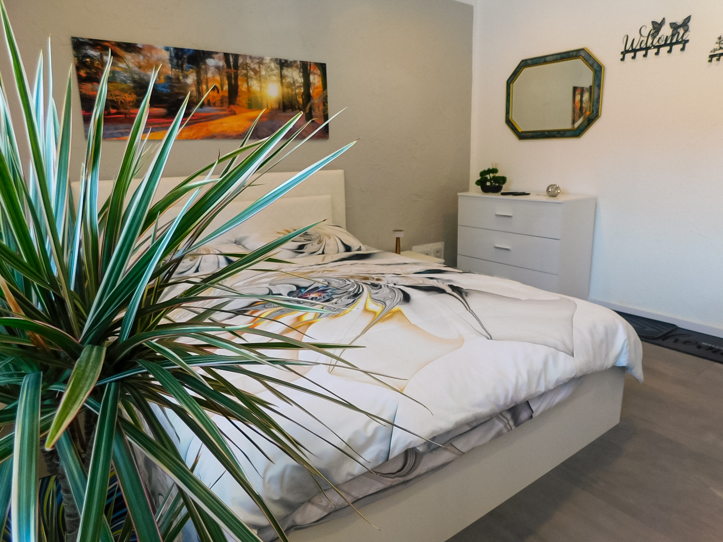 Zu sehen ist ein Schlafzimmer. Im Hintergrund ist ein großes Doppelbett zu sehen, sowie Nachttische und eine Kommode. An der Wand hängt ein Bild von einem Wald und ein Spiegel hängt über der Kommode. Im Vordergrund ist links eine Grünpflanze zu sehen. 