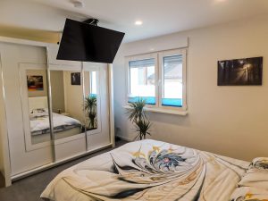 man sieht ein Bett und einen Fernseher an der Decke. Im Hintergund ist ein großer Schrank zu sehen. 