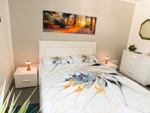 Schlafzimmer mit Doppelbett, Nachttischen, einer Kommode und einem Spiegel. An der Wand hängt ein Bild von einem Wald. 