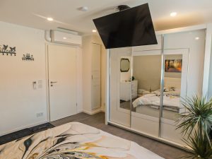 Zu sehen ist ein TV-Gerät an der Decke eines Schlafzimmers. Im Hintergrund sieht man die Zimmertür, sowie ein großer Schrank und eine grüne Pflanze. 