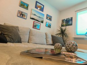 Detailaufnahme des Wohnzimmertisches. Auf dem Tisch liegen Magazine. Im Hintergrund sieht man ein Teil des Sofas, Bilder, eine grüne Pflanze und ein Fenster. 