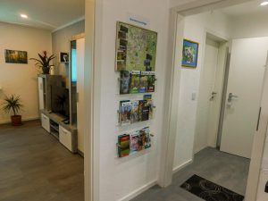 Blick in den Flur. Man sieht eine wand mit Wanderkarten in der Mitte links ist der Eingang zum Wohnzimmer zu sehen sowie die Wohnwand mit TV-gerät. Auf der rechten Seite sieht man die Eingangstür und die Tür zum Schlafzimmer. 